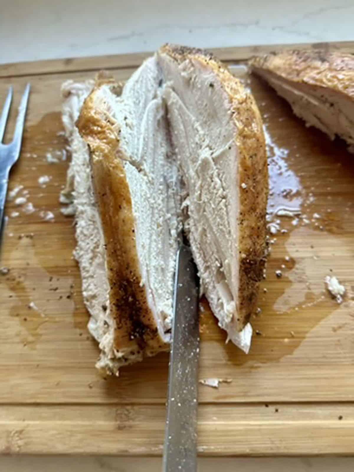 Sliced cooked chicken breast with browned seasoned skin on a wooden cutting board. A carving fork and knife are inserted into the meat, and juices are visible on the surface.