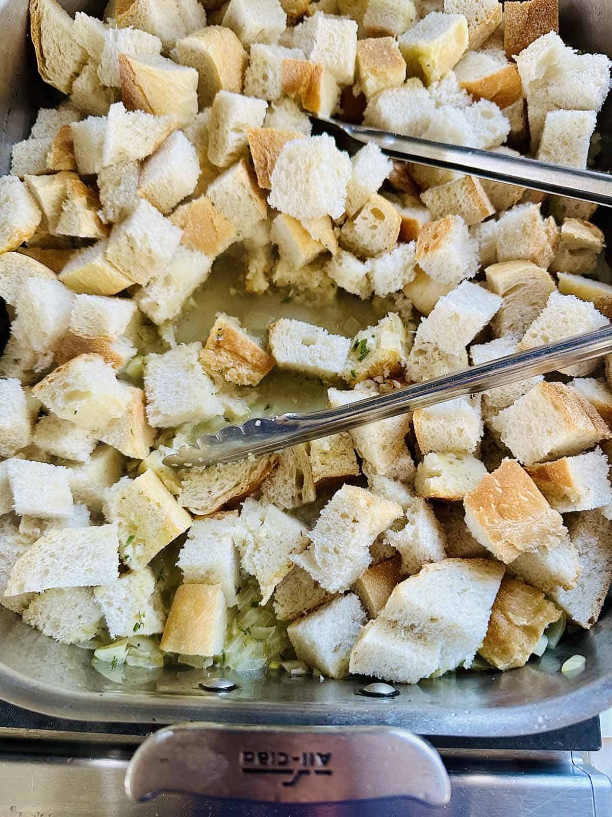 A metal pan filled with cubed bread pieces, some soaked in liquid, with metal tongs resting on top. The bread appears to be prepared for stuffing or dressing.