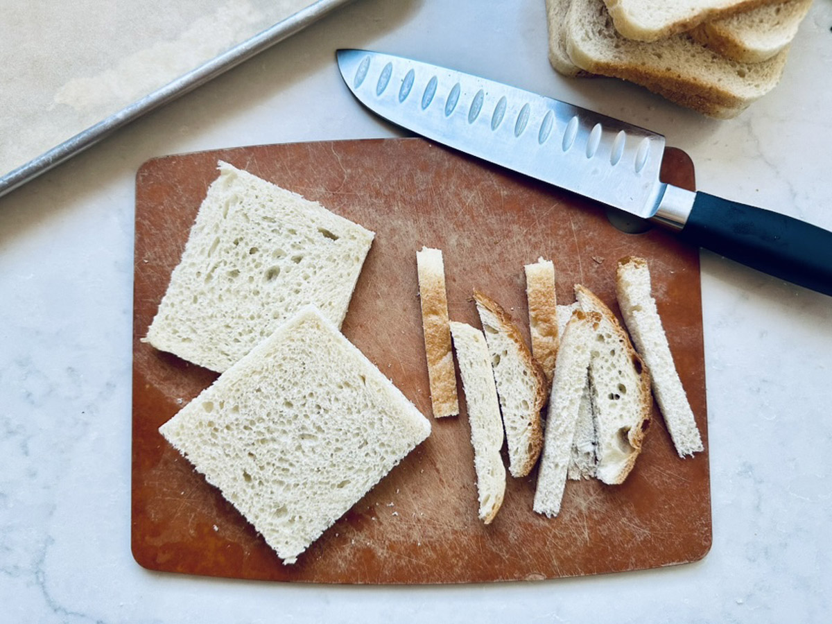 A cutting board with two square slices of white bread, several bread crust strips, a chef’s knife, and a stack of bread slices in the background on a white surface.