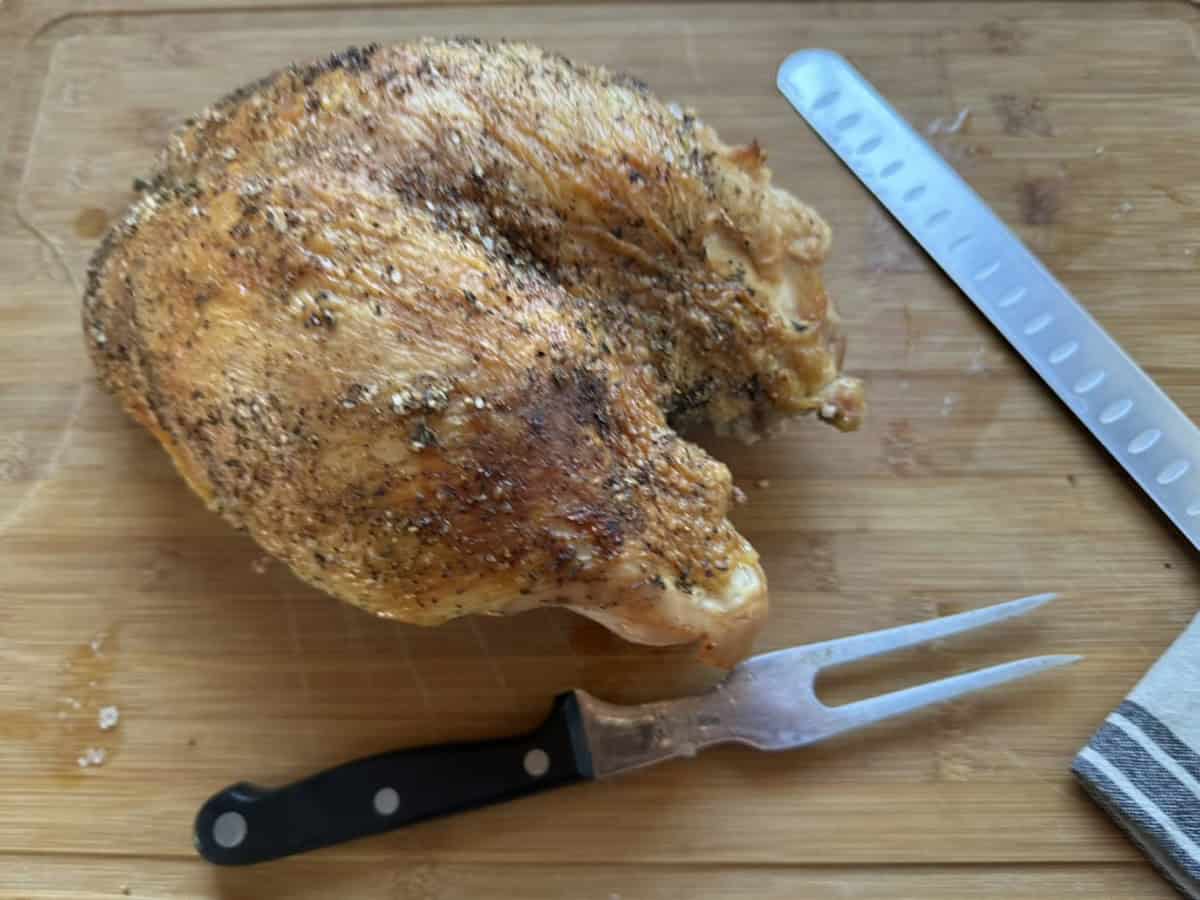 A roasted, seasoned chicken rests on a wooden cutting board next to a carving fork and a slicing knife. A striped kitchen towel is partially visible in the corner.