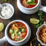 Two bowls of tortilla soup topped with avocado slices, jalapeño, cheese, and tortilla strips on a wooden table, surrounded by lime wedges, cilantro, sour cream, and extra tortilla strips.