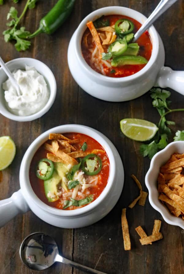 Two bowls of tortilla soup topped with avocado slices, jalapeño, cheese, and tortilla strips on a wooden table, surrounded by lime wedges, cilantro, sour cream, and extra tortilla strips.