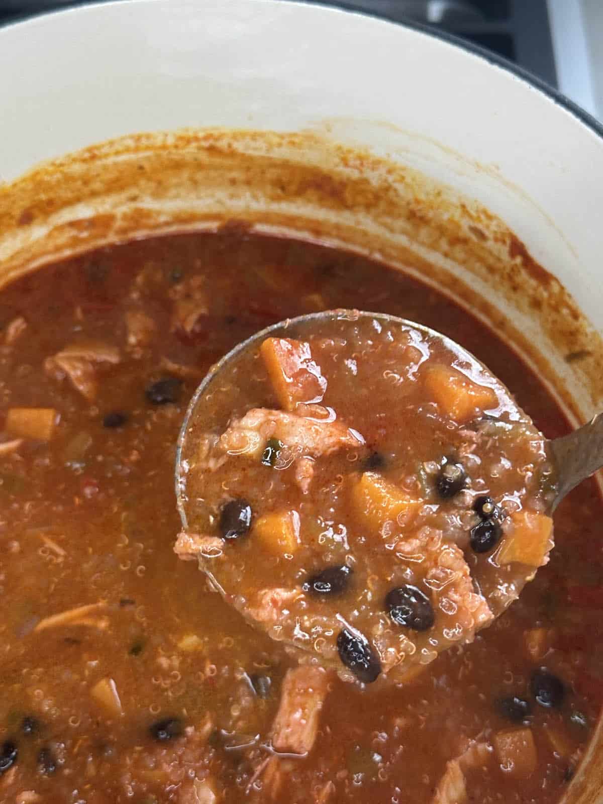 A close-up of a pot filled with hearty chili, featuring black beans, chunks of tomato, carrot, and shredded meat being scooped up with a ladle.