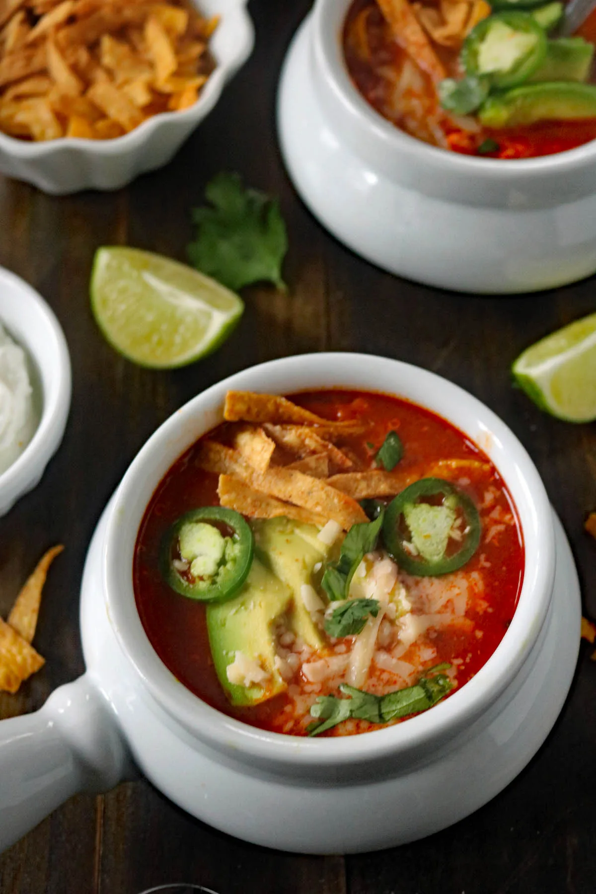 A bowl of tortilla soup topped with avocado slices, jalapeño, shredded cheese, crispy tortilla strips, and cilantro, with lime wedges and a bowl of sour cream on a dark wooden table.