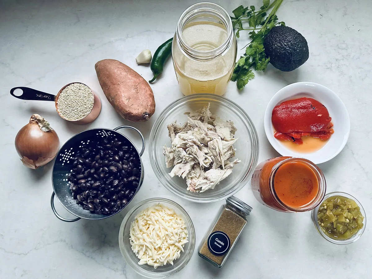 A flat lay of various ingredients on a white surface, including shredded chicken, black beans, sweet potato, cheese, onion, jar of broth, red pepper, green chilies, quinoa, sauce, an avocado, and fresh herbs.