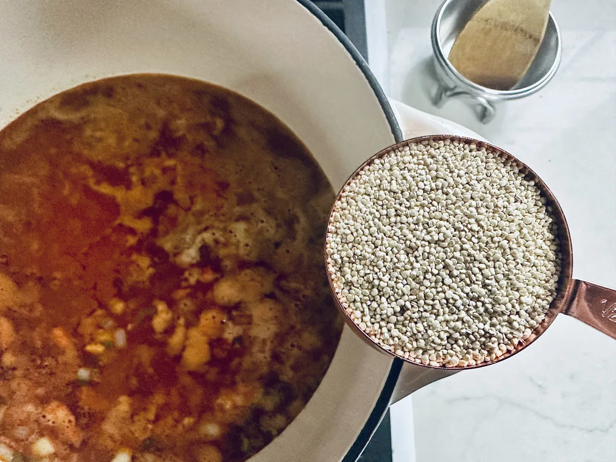 A measuring cup filled with uncooked quinoa is held above a pot of simmering soup or stew on a stovetop, with a wooden spoon resting nearby.