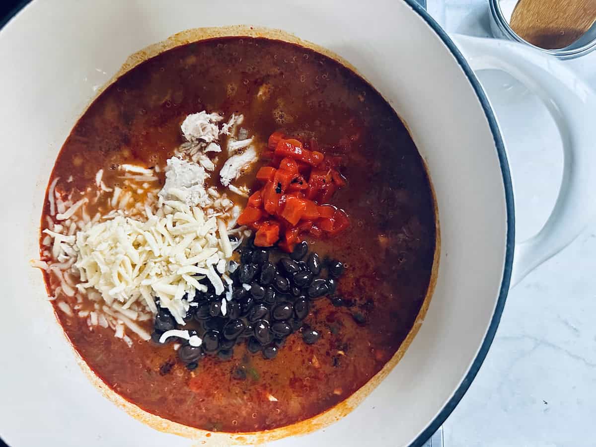 A white pot on a stovetop filled with chili, topped with shredded cheese, black beans, diced red peppers, and a dollop of sour cream before mixing.