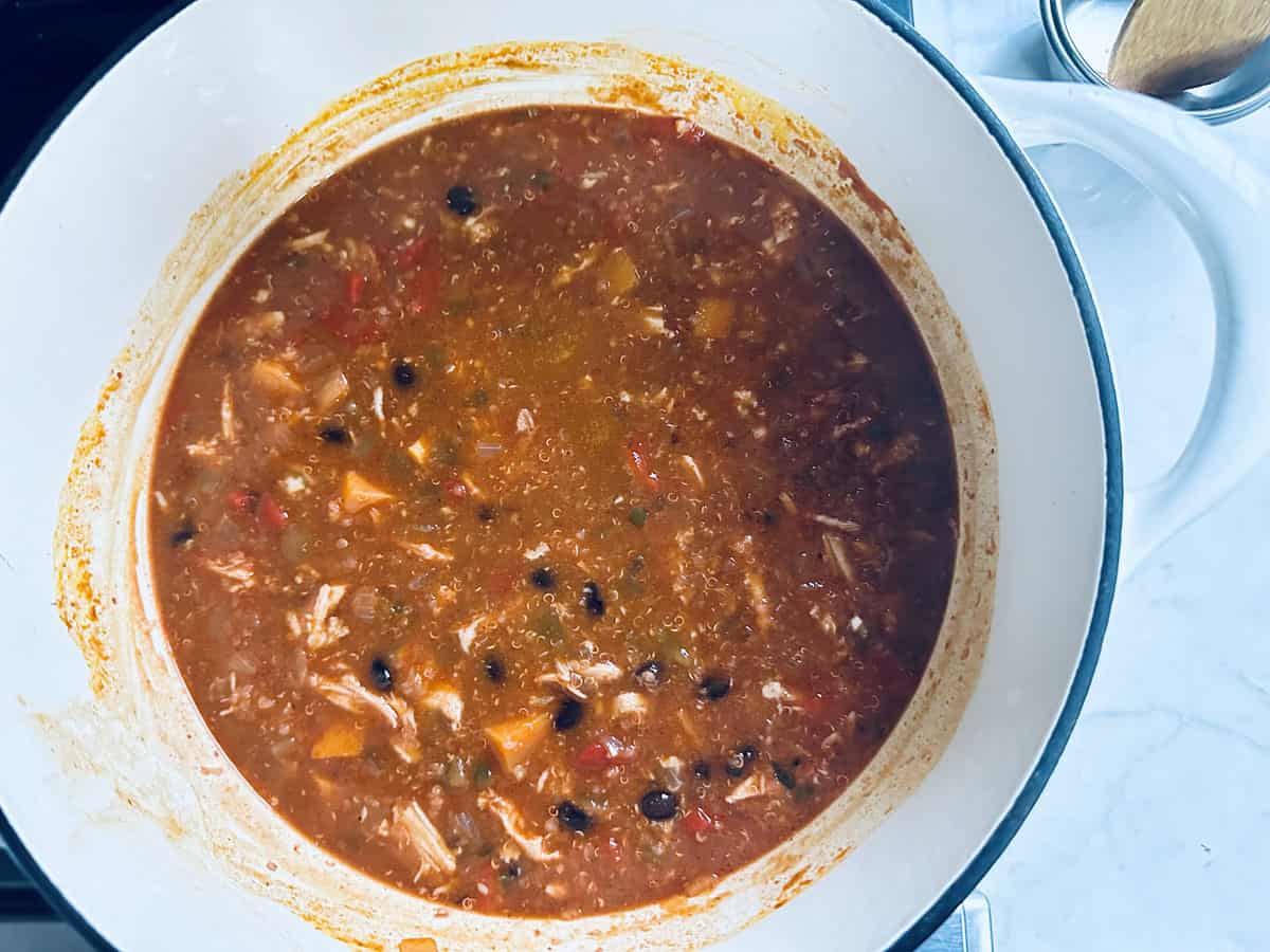 A large white pot on a stove is filled with a hearty stew containing shredded meat, black beans, diced tomatoes, and chunks of yellow and orange vegetables in a rich, reddish broth.