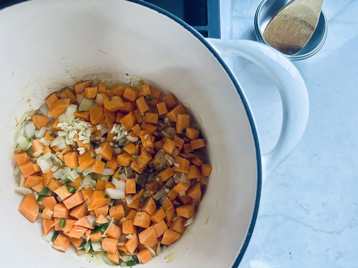 A white pot on a stove contains chopped onions, carrots, and sweet potatoes being sautéed. A small metal bowl and a wooden spoon rest on a white countertop nearby.