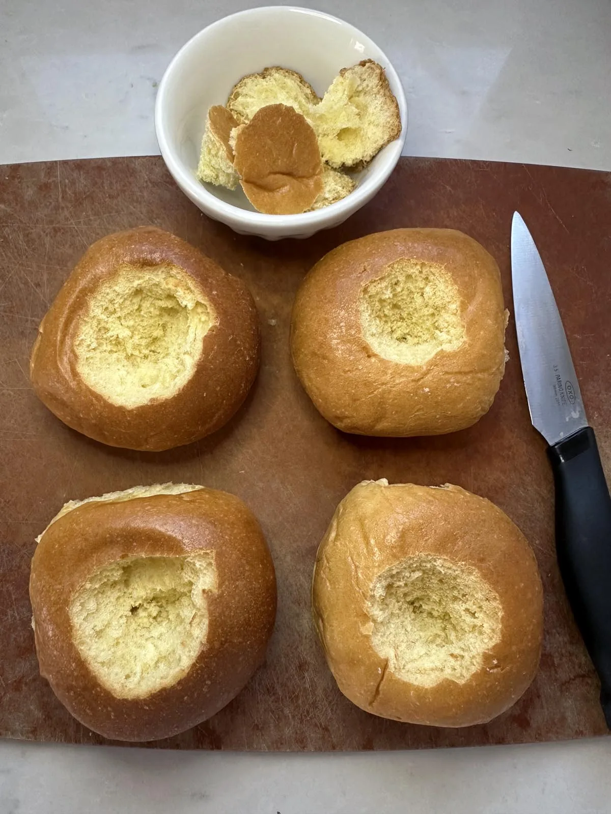 Four bread rolls with their tops hollowed out sit on a brown cutting board next to a knife, with the removed bread pieces placed in a small white bowl.