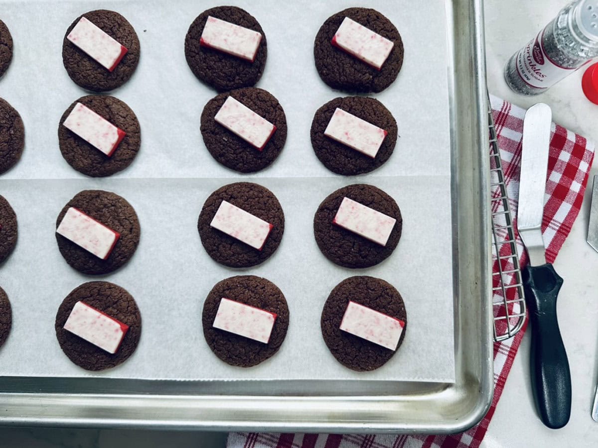 Rows of chocolate cookies on a baking sheet, each topped with a rectangular piece of peppermint bark. A red and white kitchen towel and a spatula are visible to the side.