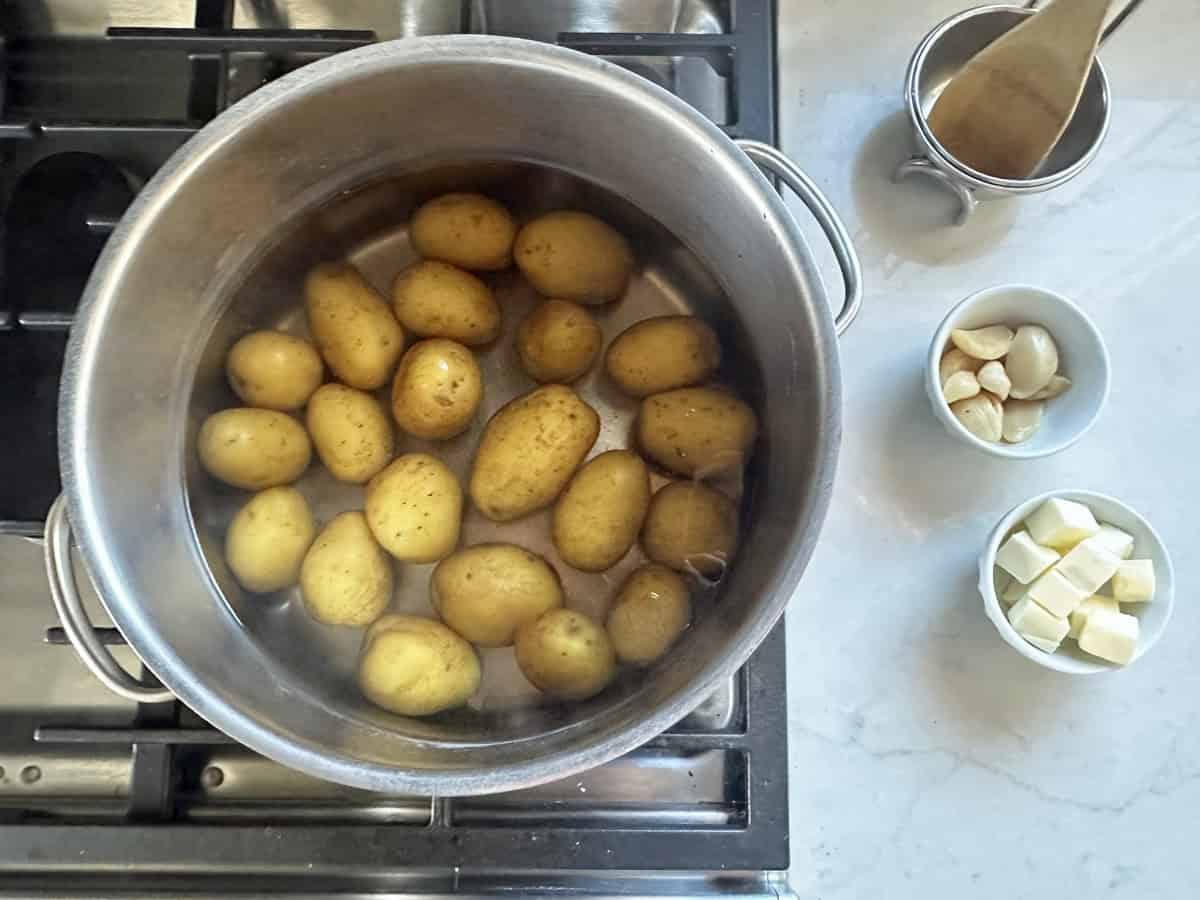 Top view of a pot of whole potatoes boiling on a stove. On the counter beside the pot are a wooden spoon in a cup, a bowl of garlic cloves, and a bowl of butter cubes.