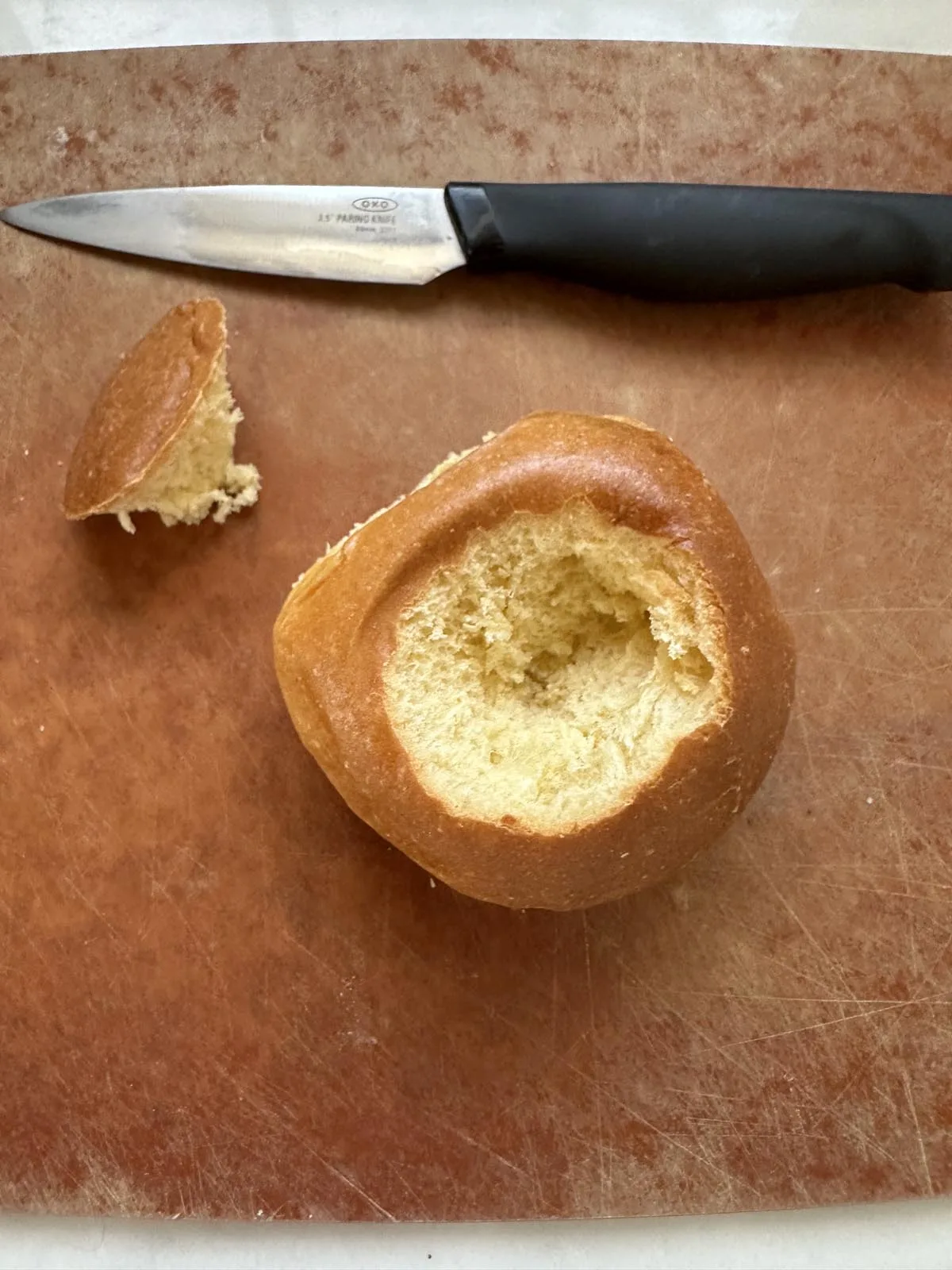 A bread roll with the top cut off and the inside hollowed out sits on a brown cutting board next to a knife with a black handle.