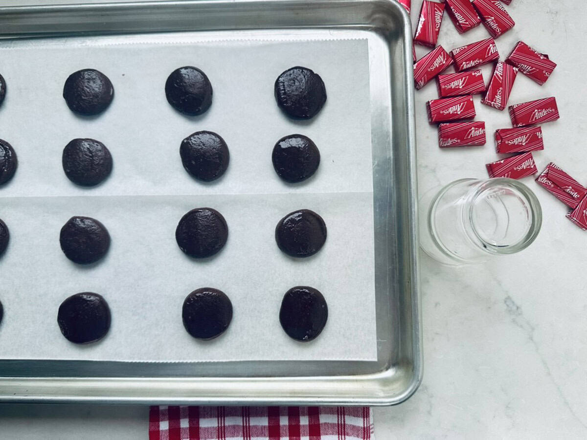 A baking sheet lined with parchment paper holds rows of unbaked chocolate cookie dough balls. To the right, red-wrapped Andes mints and a small empty glass jar sit on a white countertop with a red-striped cloth.