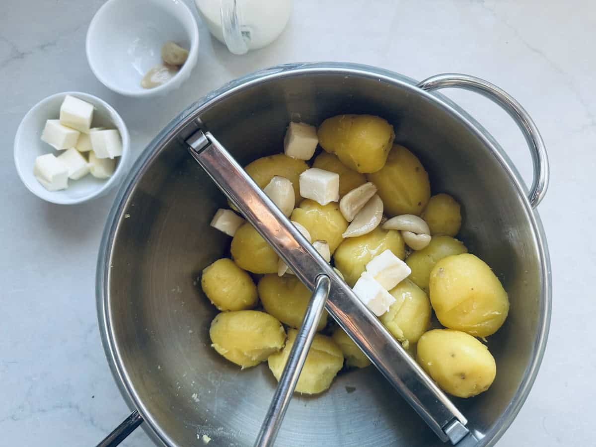 A metal pot filled with peeled boiled potatoes, garlic cloves, and butter cubes, with a food mill placed on top. Two small bowls nearby hold more butter cubes and garlic.