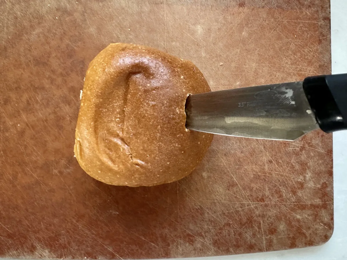 A serrated knife is slicing through a small, brown bread roll placed on a worn, reddish-brown cutting board.