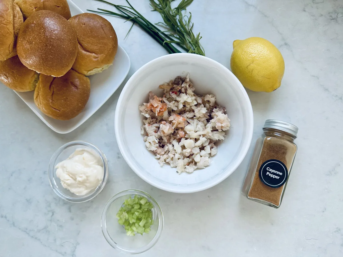 A bowl of chopped seafood sits on a white countertop, surrounded by slider buns, fresh herbs, a lemon, a jar of cayenne pepper, a small bowl of mayonnaise, and a small bowl of chopped celery.