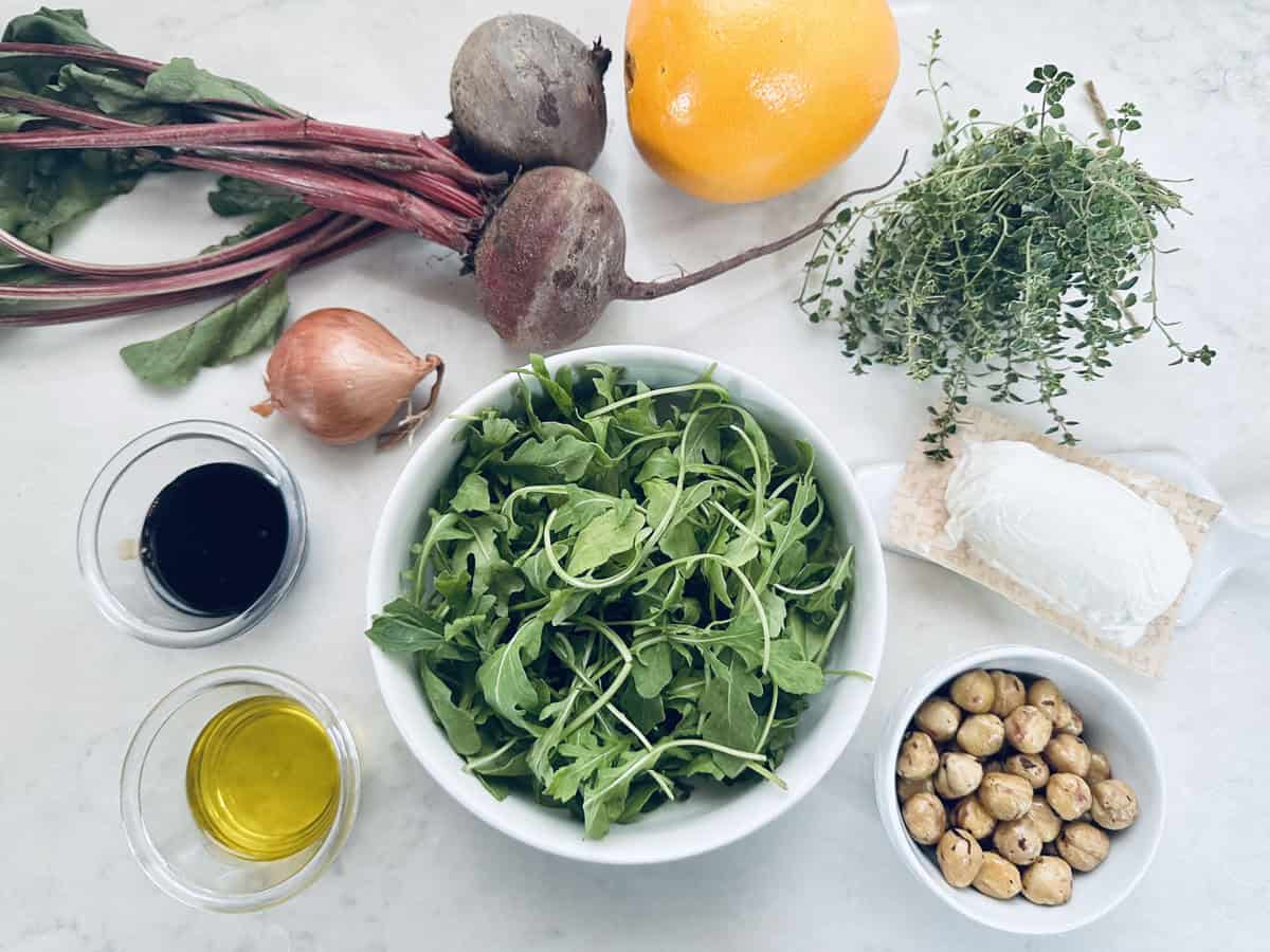 A bowl of arugula is surrounded by beets, a lemon, fresh thyme, a shallot, goat cheese, roasted chickpeas, olive oil, and balsamic vinegar on a white surface.