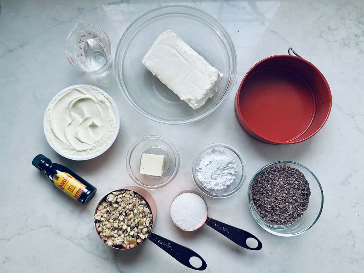 A top-down view of various baking ingredients in bowls and cups, including cream cheese, sour cream, butter, powdered sugar, chopped nuts, chocolate shavings, vanilla extract, granulated sugar, water, and a red baking pan.