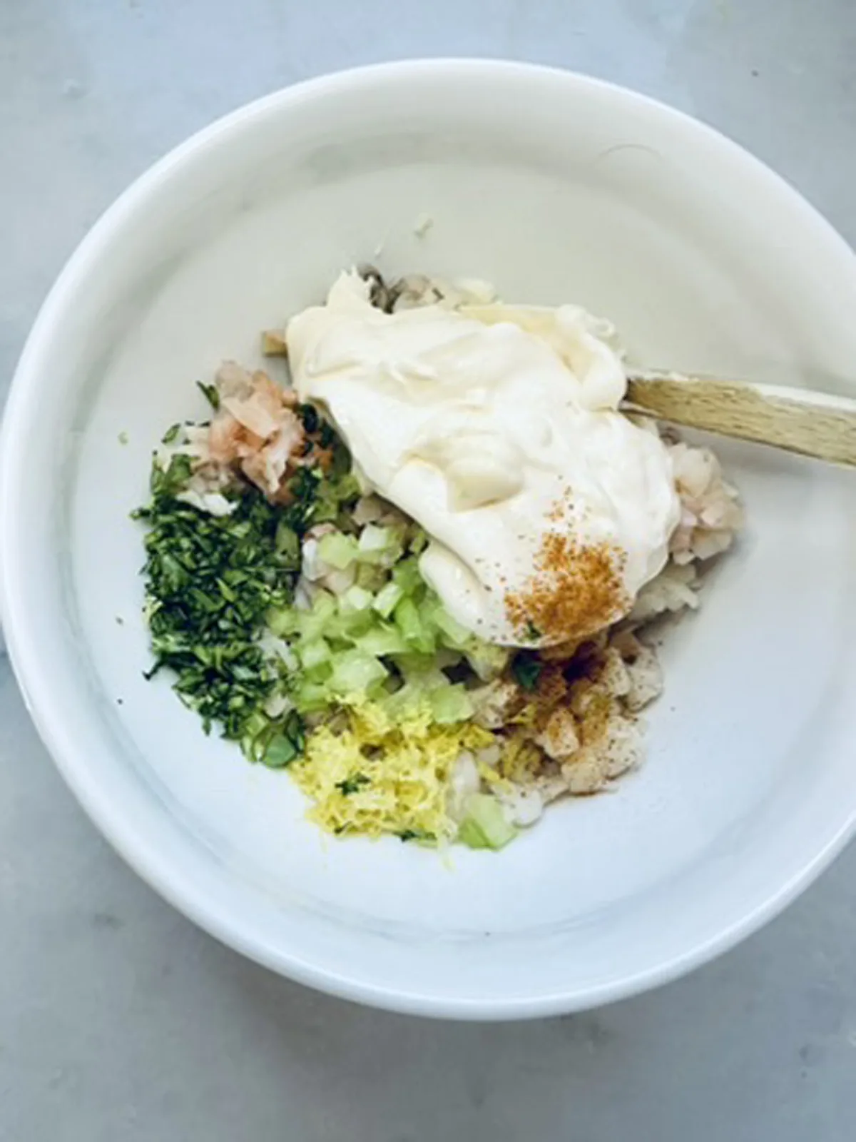 A white bowl contains chopped herbs, diced celery, onion, lemon zest, seasonings, and a large dollop of mayonnaise, ready to be mixed with a wooden spoon on a light surface.