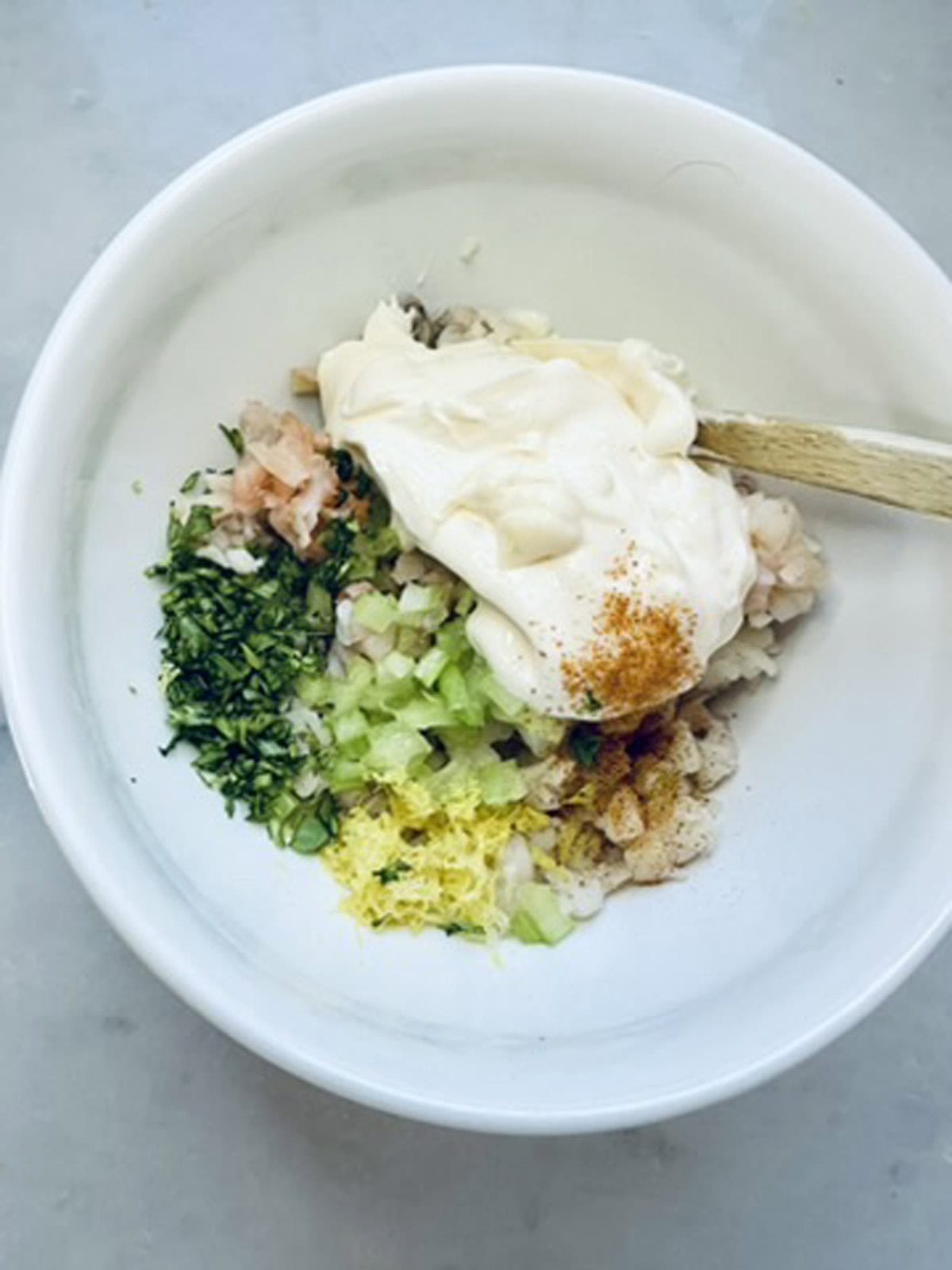 A white bowl contains chopped herbs, diced celery, onion, lemon zest, seasonings, and a large dollop of mayonnaise, ready to be mixed with a wooden spoon on a light surface.