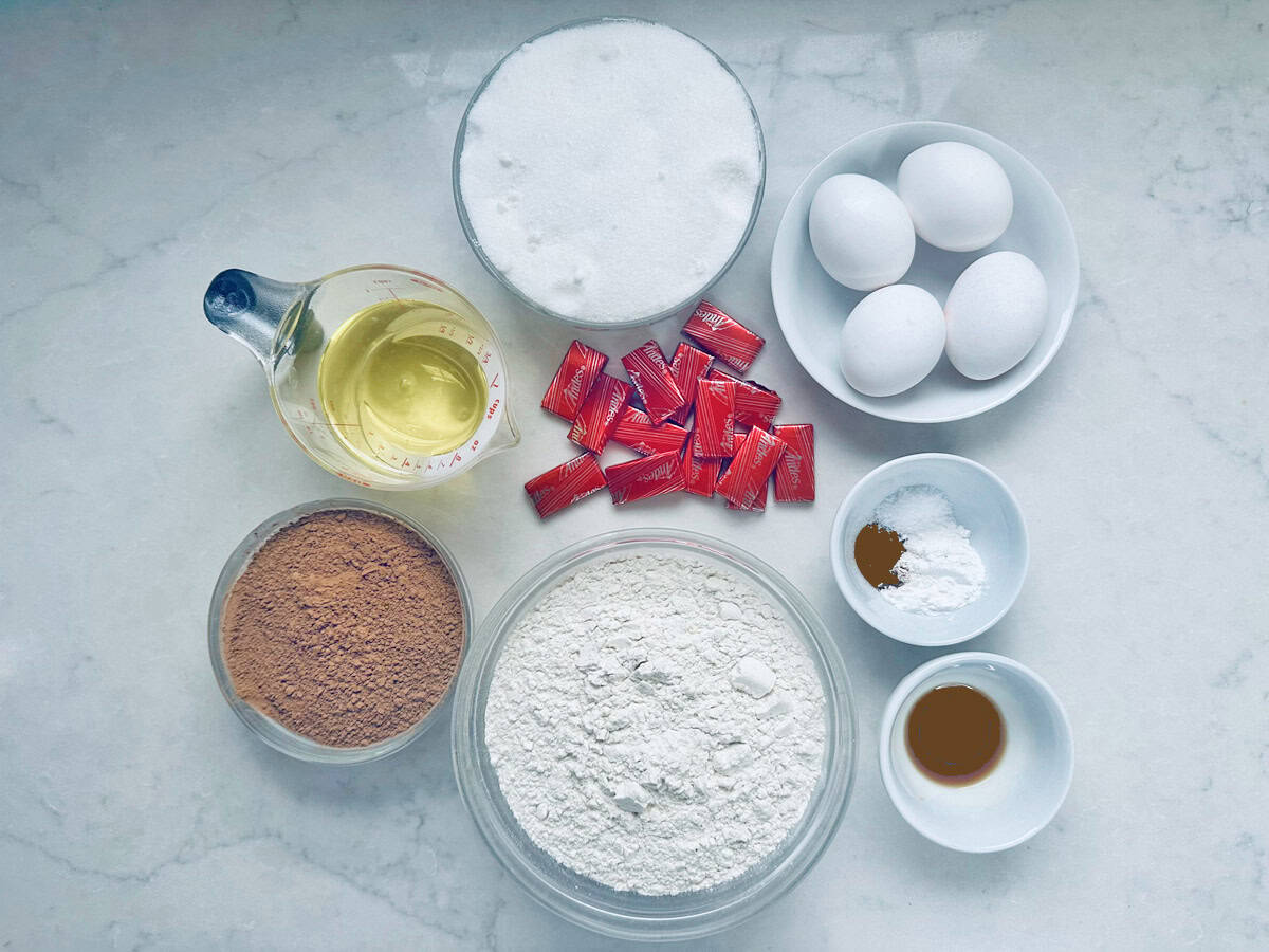 Baking ingredients on a counter: eggs, granulated sugar, cooking oil, flour, cocoa powder, red food coloring packets, baking powder, vanilla extract, and cinnamon, all in separate bowls and containers.