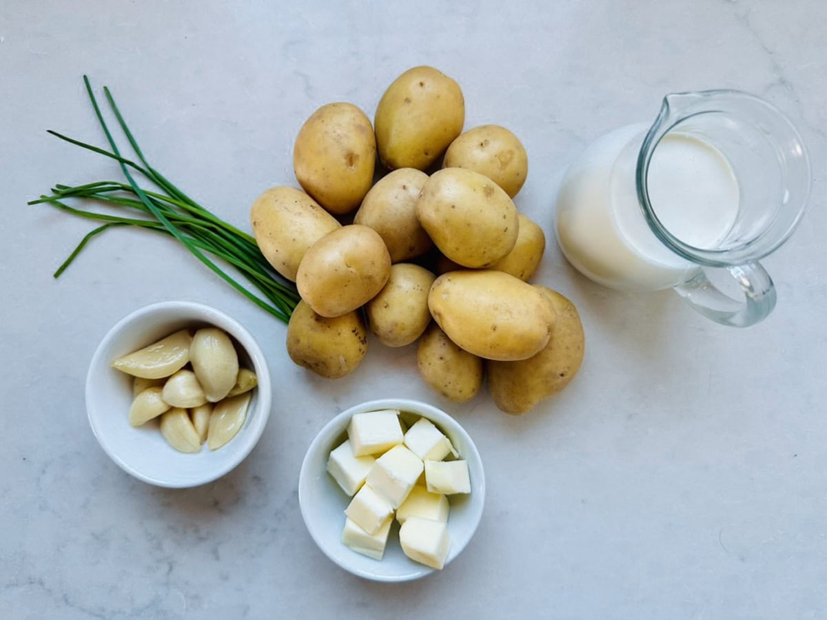 Several whole potatoes, a bunch of chives, a small bowl of peeled garlic cloves, a bowl of cubed butter, and a glass pitcher of milk arranged on a white surface.