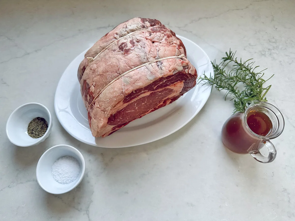 A Standing Rib Roast with Au Jus sits on a white plate, tied with string. Beside it are a sprig of rosemary, a small jug of savory au jus, and two small bowls containing salt and pepper.