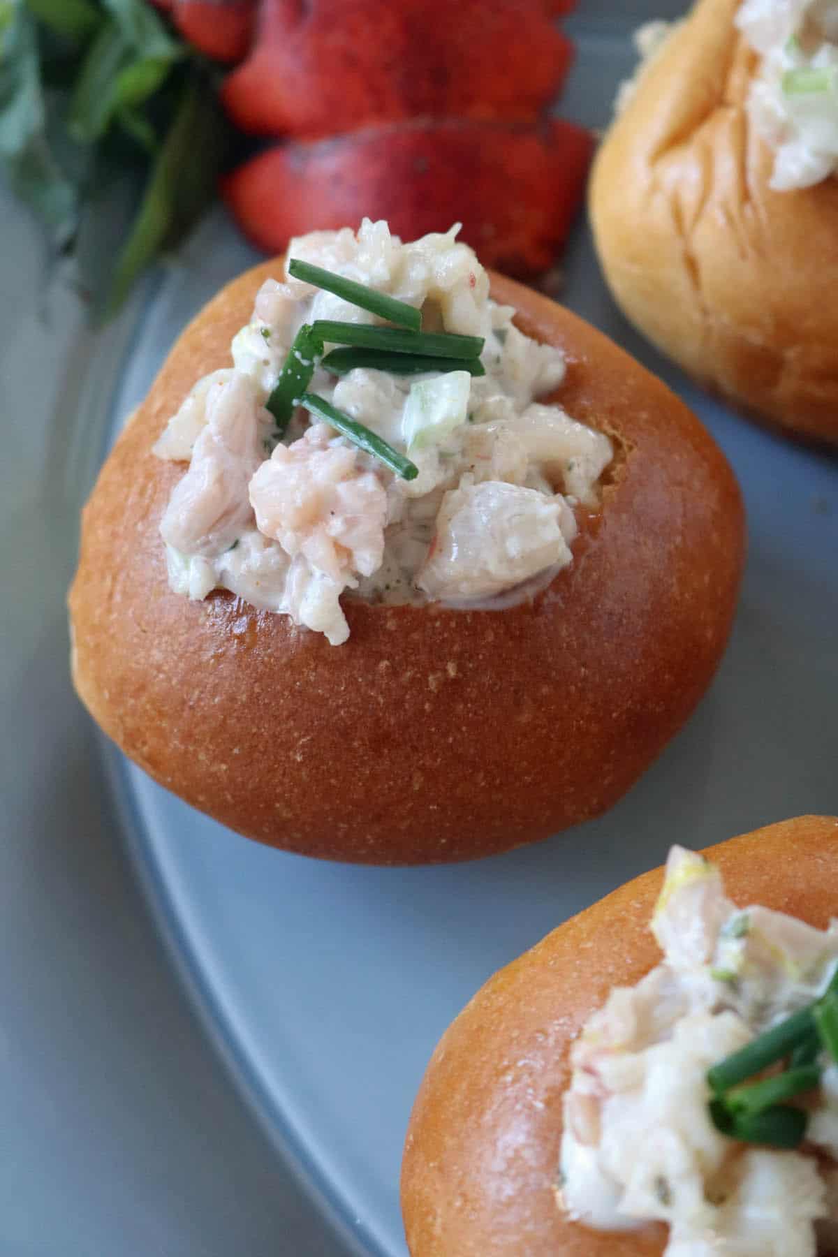 A close-up of a bread roll filled with creamy lobster salad, garnished with chopped chives, on a gray plate.