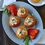 A platter with five bread rolls stuffed with lobster salad, garnished with chives, and flanked by two lobster tails. A small plate with tarragon, napkins, and lobster is nearby on a wooden table.