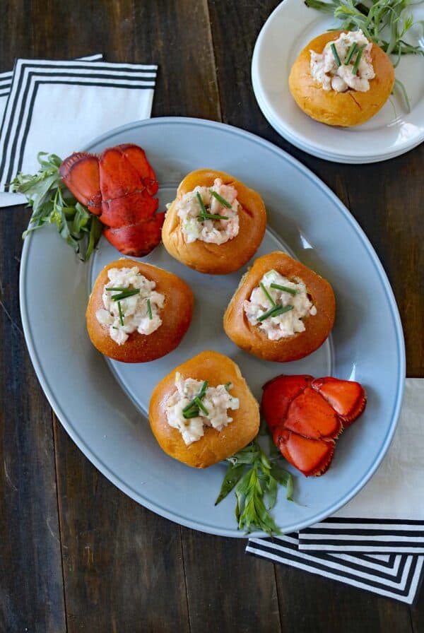 A platter with five bread rolls stuffed with lobster salad, garnished with chives, and flanked by two lobster tails. A small plate with tarragon, napkins, and lobster is nearby on a wooden table.