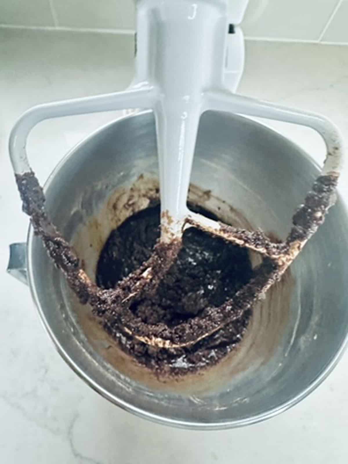 A stand mixer with a paddle attachment is mixing dark brown batter in a metal bowl, set on a light-colored countertop.