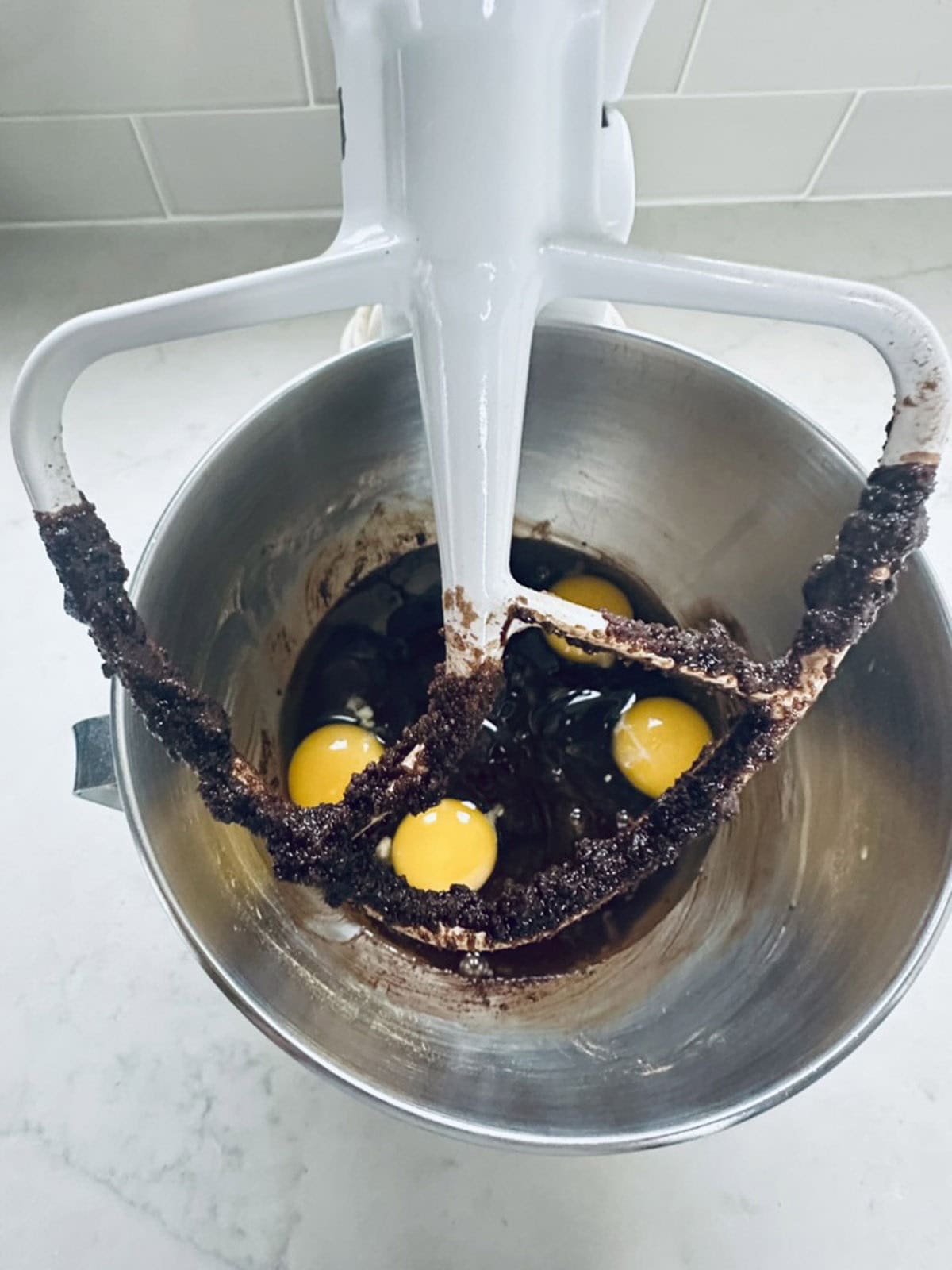 A mixing bowl with brownie batter, three raw eggs, and a mixer paddle attachment coated with chocolate resting above the bowl.
