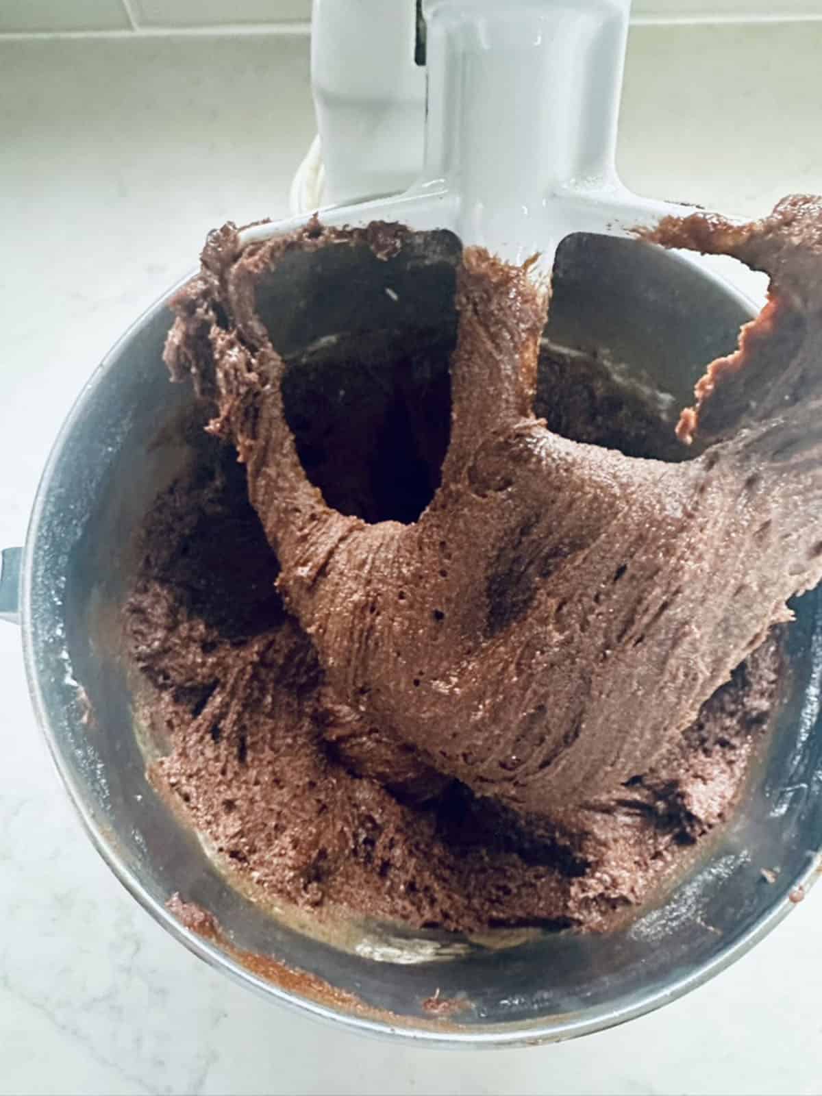 A stand mixer bowl filled with chocolate batter, with some of the batter clinging to the attached paddle mixer on a light-colored countertop.