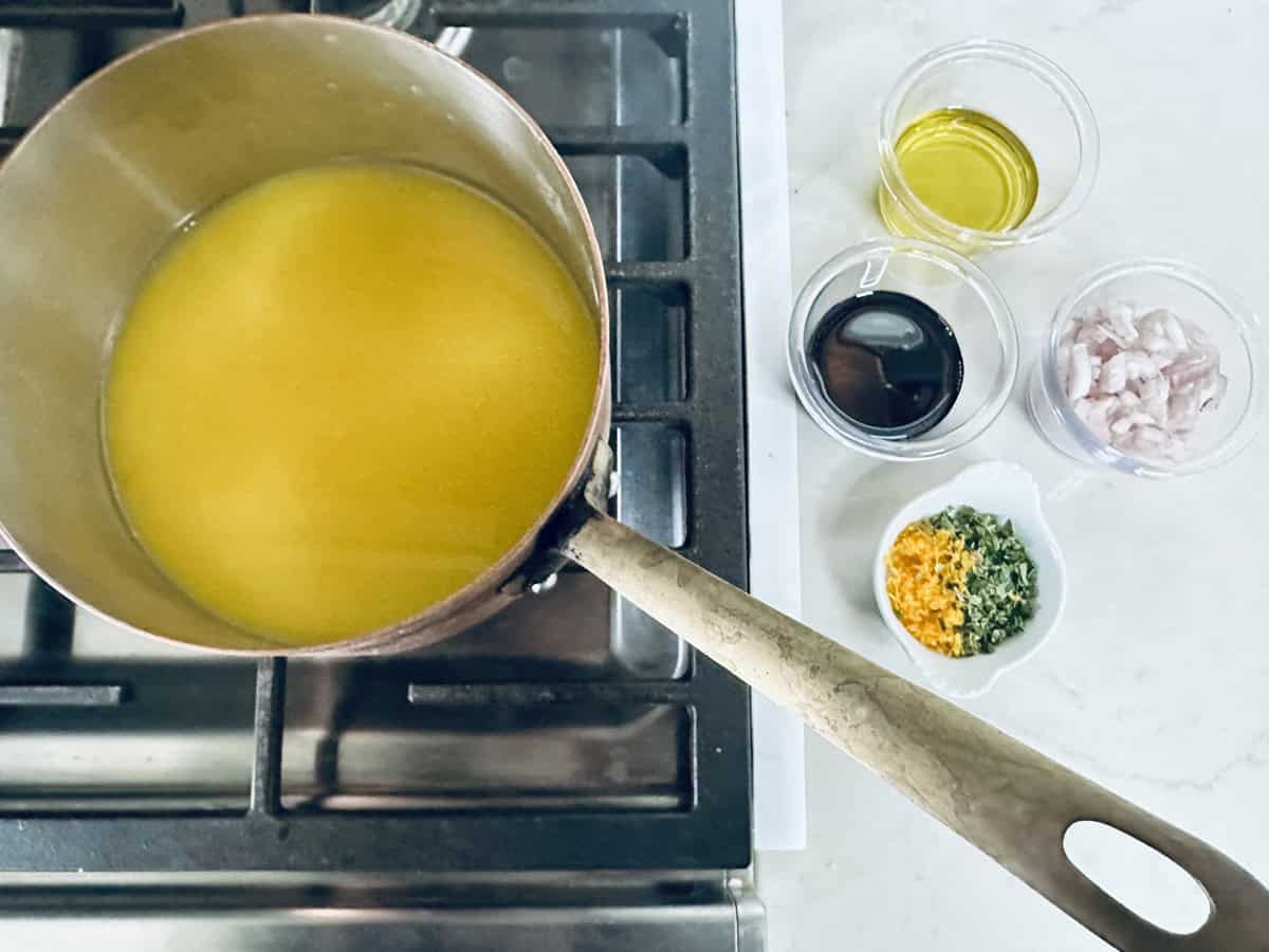 A saucepan with yellow liquid is on a stove. Next to it on the counter are small bowls containing olive oil, balsamic vinegar, chopped shallots, orange zest, and green herbs.