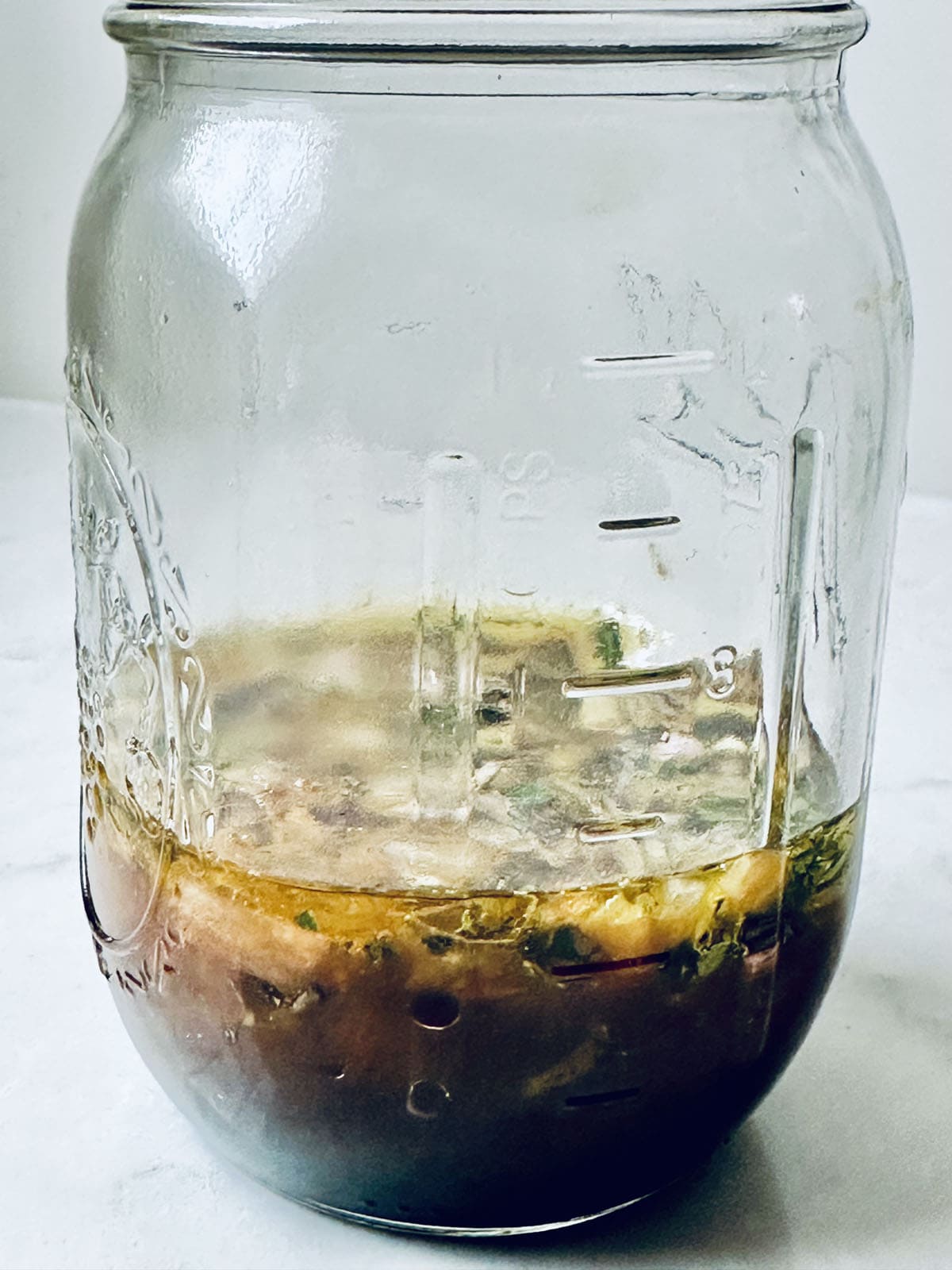 A large glass mason jar with measurement markings containing a small amount of brown liquid and chopped herbs at the bottom, sitting on a white surface.