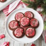 A plate of chocolate cookies topped with red and white swirled frosting and sprinkles sits on a wooden table next to a holiday towel, evergreen branches, and several unwrapped Andes mints. More cookies are on a tray in the background.