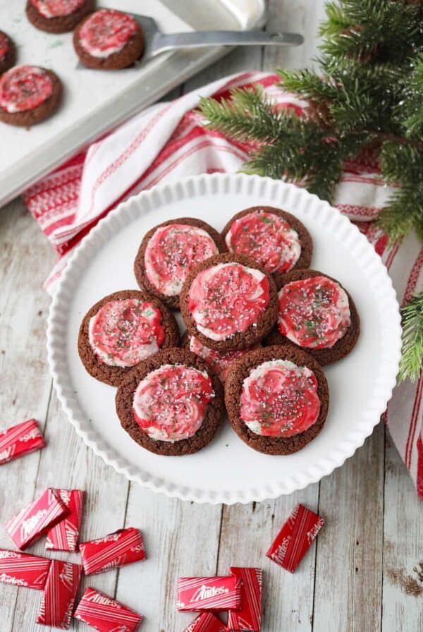 A plate of chocolate cookies topped with red and white swirled frosting and sprinkles sits on a wooden table next to a holiday towel, evergreen branches, and several unwrapped Andes mints. More cookies are on a tray in the background.