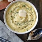 A bowl of creamy mashed potatoes topped with a pat of butter and chopped chives, placed on a wooden table next to a spoon, garlic, and a small dish of chopped herbs.