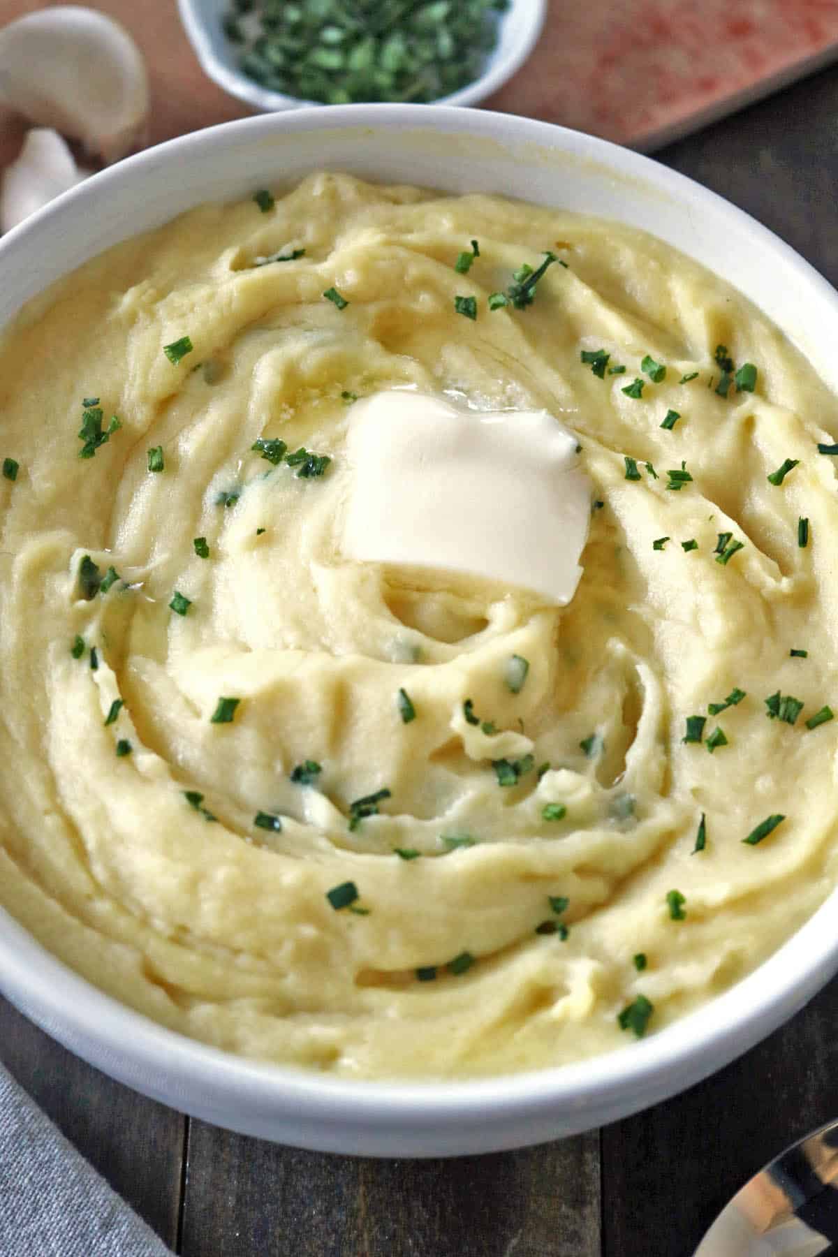 A bowl of creamy mashed potatoes topped with a pat of melting butter and sprinkled with chopped chives.