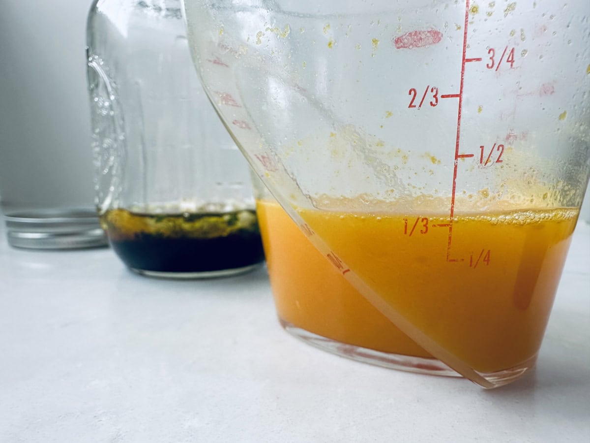 A glass measuring cup with orange liquid sits on a countertop, showing measurements on the side. Behind it, there is a jar containing a dark liquid. The background is softly blurred.