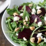 A white bowl filled with arugula, roasted beet slices, crumbled goat cheese, and chopped nuts sits on a dark wooden table with a napkin and two forks in the background.