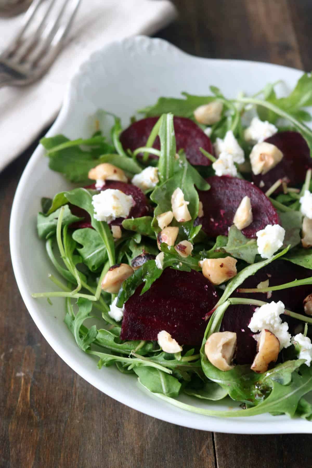 A white bowl filled with arugula, roasted beet slices, crumbled goat cheese, and chopped nuts sits on a dark wooden table with a napkin and two forks in the background.
