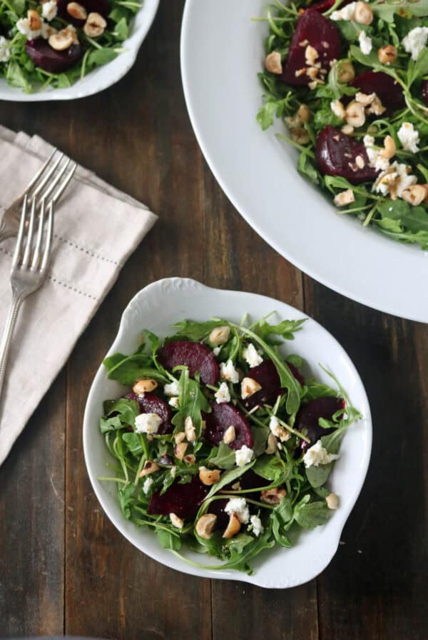 A bowl of arugula salad with roasted beets, crumbled cheese, and chopped nuts sits on a wooden table near a napkin with two forks and a larger dish of the same salad.