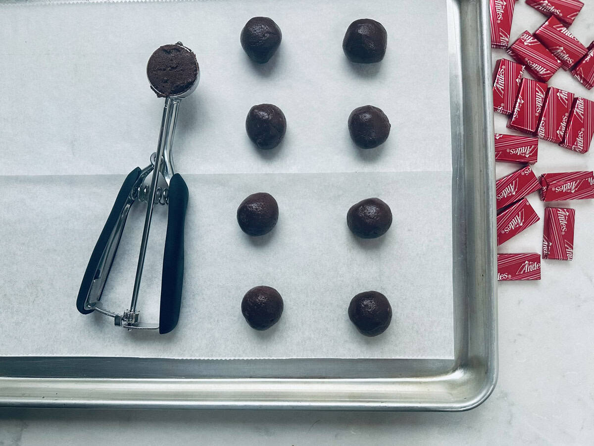 A baking sheet with eight evenly spaced chocolate dough balls on parchment paper, a cookie scoop with dough, and a pile of red-wrapped Andes mints on the side.