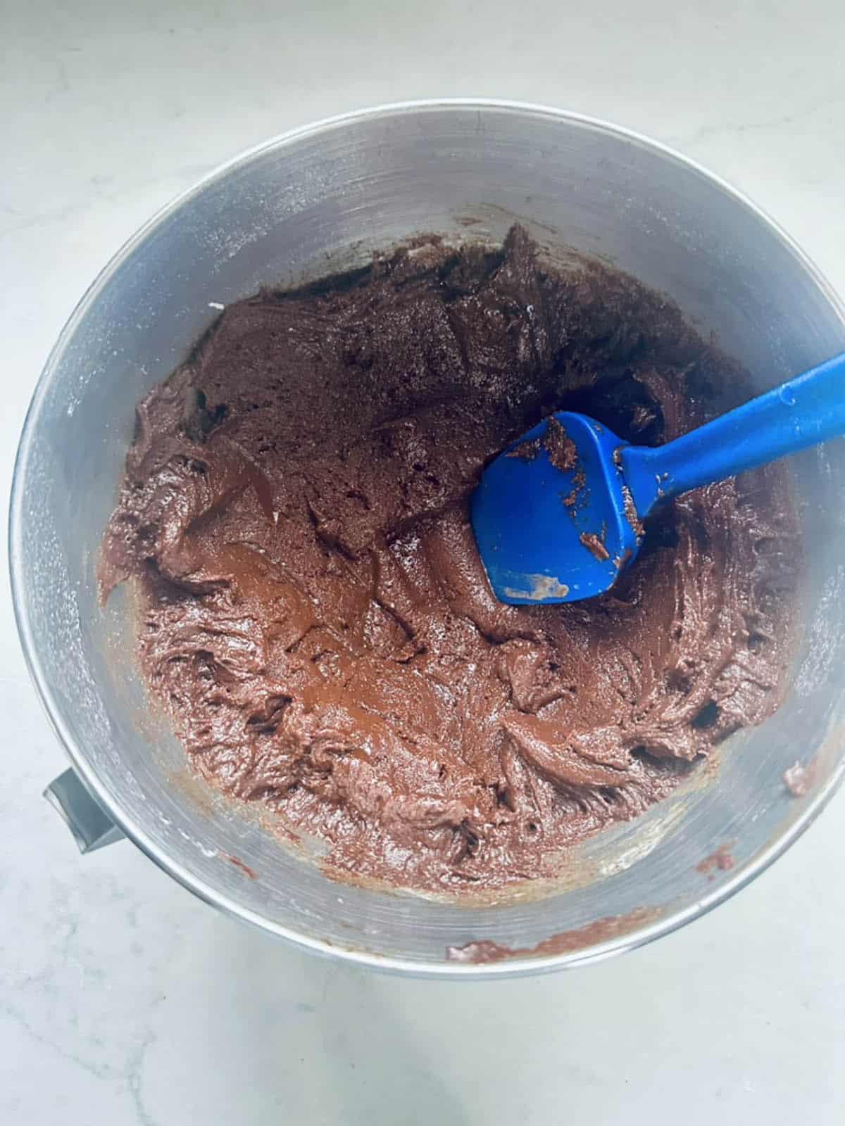 A metal mixing bowl filled with chocolate batter, being stirred with a blue spatula, sits on a light-colored countertop.