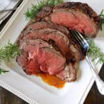 A white platter with slices of medium-rare Standing Rib Roast with Au Jus, garnished with sprigs of fresh rosemary. A serving fork rests beside the meat, and a small dish of sauce is in the background.