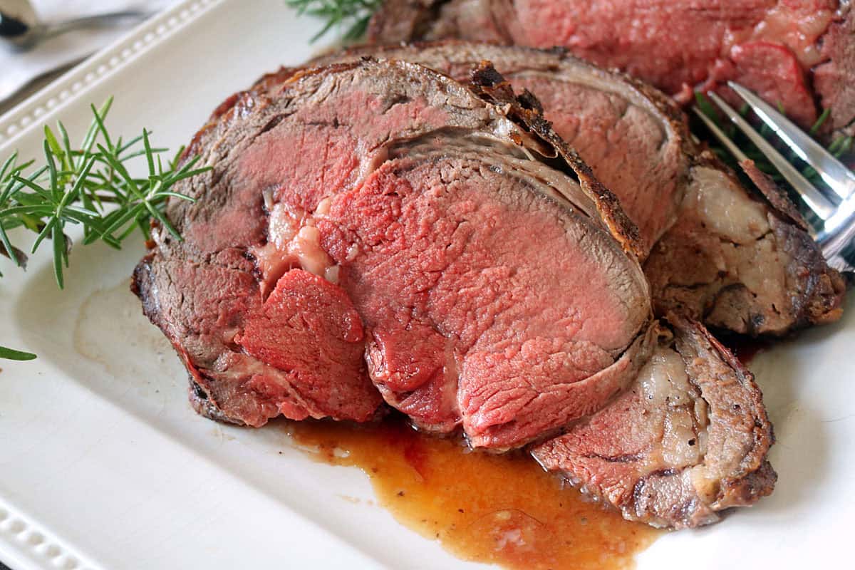 Two thick slices of medium-rare Standing Rib Roast with Au Jus sit on a white plate, garnished with fresh rosemary sprigs, savory juices pooling underneath and a fork visible to the side.