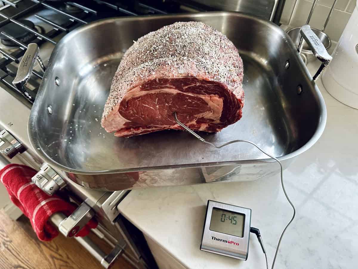 A seasoned Standing Rib Roast with Au Jus sits in a roasting pan, a meat thermometer probe inserted, displaying a timer reading 0:45 on the kitchen counter near the stove.