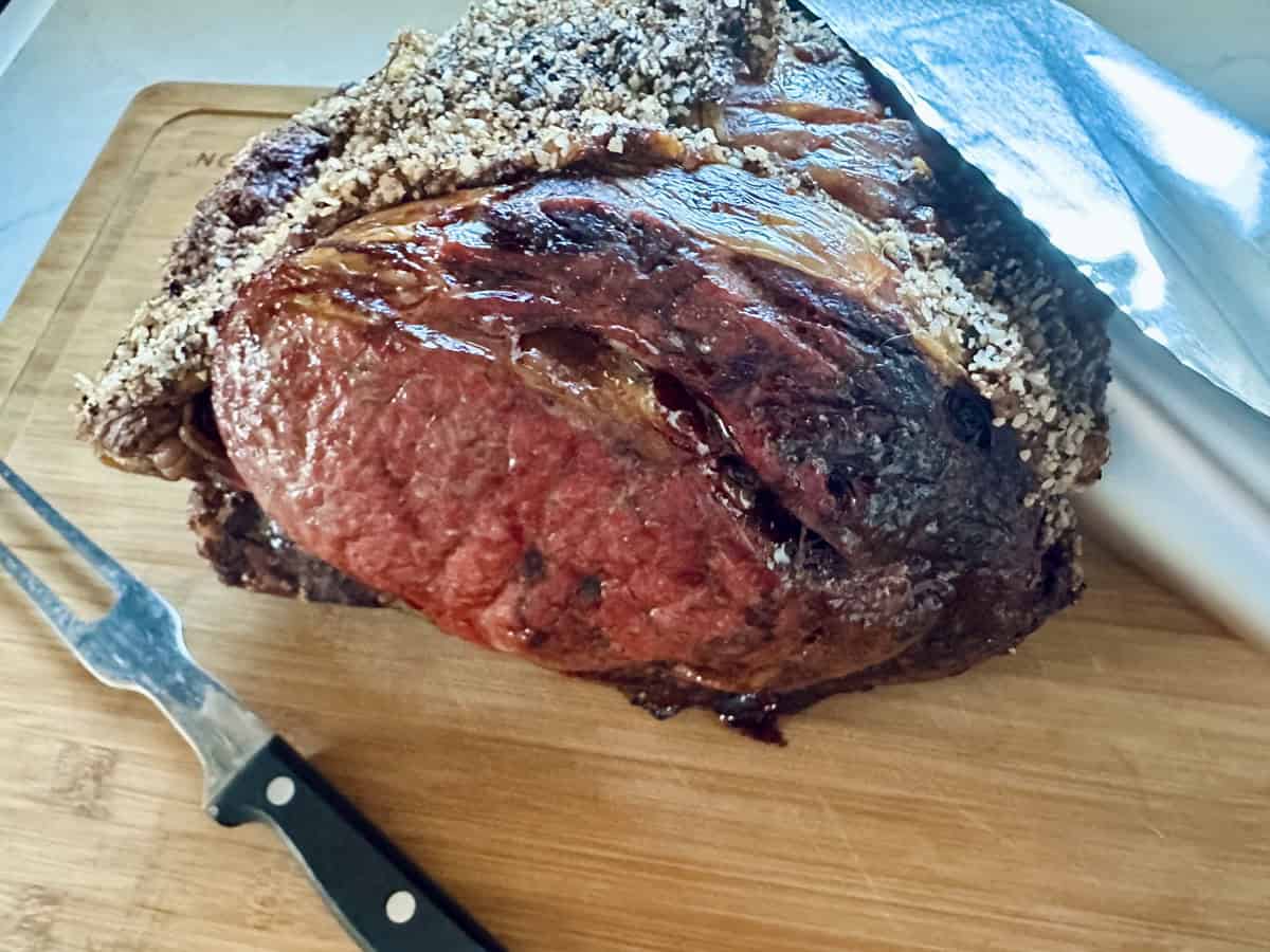 A cooked Standing Rib Roast with Au Jus, boasting a crispy, seasoned crust, rests on a wooden cutting board beside a carving fork and a large knife.